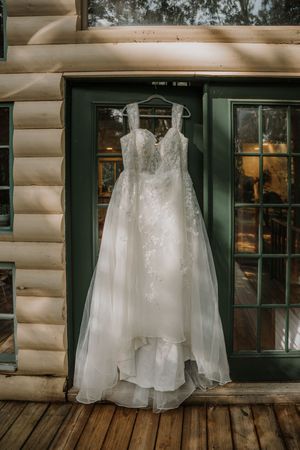 Elegant lace wedding gown with floral appliques hanging on a green door of a rustic log cabin.