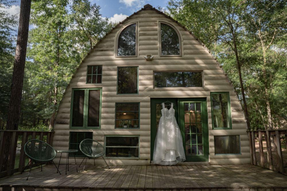 White wedding dress hanging on green doors of a rustic log cabin surrounded by lush green trees.