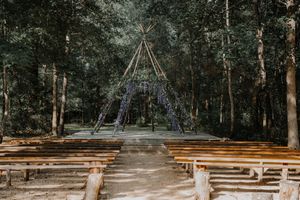 Rustic outdoor wedding ceremony setup with wooden benches and a teepee arch decorated with purple flowers.