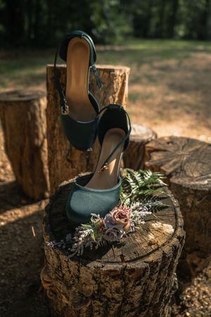 Teal bridal heels displayed on a tree stump with dried floral boutonniere in a sunlit forest setting.