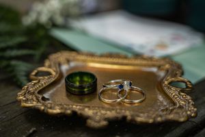 Gold wedding rings and a green groom band arranged on an ornate vintage gold tray with greenery.
