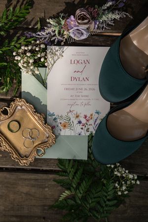 Wedding flat lay featuring floral invitation for Logan and Dylan, teal heels, rings, and wildflower bouquet.