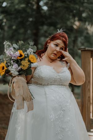 Emotional red-haired bride in lace gown wiping tears while holding a vibrant sunflower and purple bouquet.