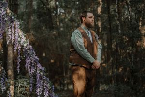 Groom in brown vest and sage green shirt stands beside purple wisteria flowers in a woodland setting.
