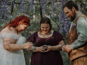 Bride and groom exchange rings during an outdoor forest wedding ceremony with purple wisteria flowers.