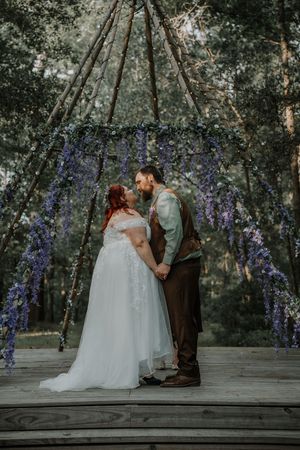 Newlyweds share a kiss under a rustic teepee arch adorned with cascading purple wisteria in a forest.