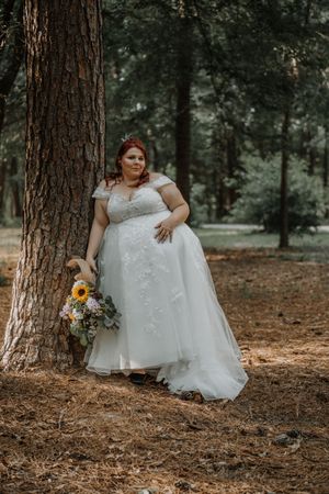 Plus-size bride in lace off-shoulder gown leaning against pine tree holding sunflower bouquet.