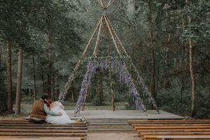 Newlyweds sit together on a wooden stage beneath a wisteria-draped teepee arch in a forest.