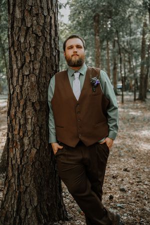 Dapper groom in brown vest and sage shirt leans against a pine tree in a forest setting.