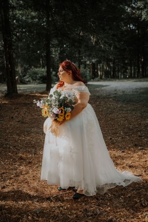 Bride in white off-shoulder wedding gown holding colorful bouquet standing in sunlit forest.