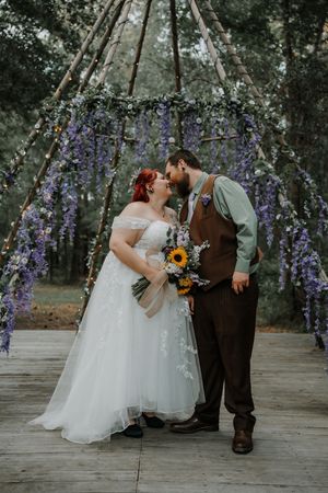 Bride and groom kiss holding a sunflower bouquet beneath a wisteria-covered teepee arch in a forest.