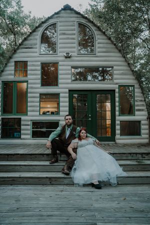 Bride and groom posing on steps of rustic log cabin venue surrounded by trees.