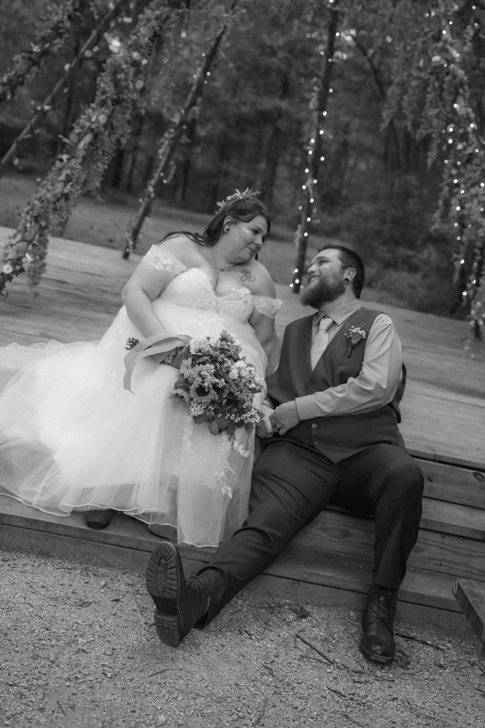 Black and white photo of bride and groom sitting on wooden platform under string lights.