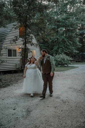 Bride and groom walking hand in hand on gravel path beside log cabin with string lights.