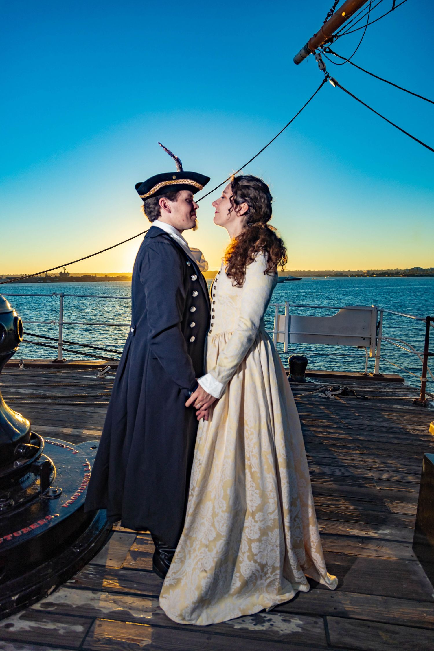 Couple in period costumes sharing a romantic moment on a tall ship deck at sunset.