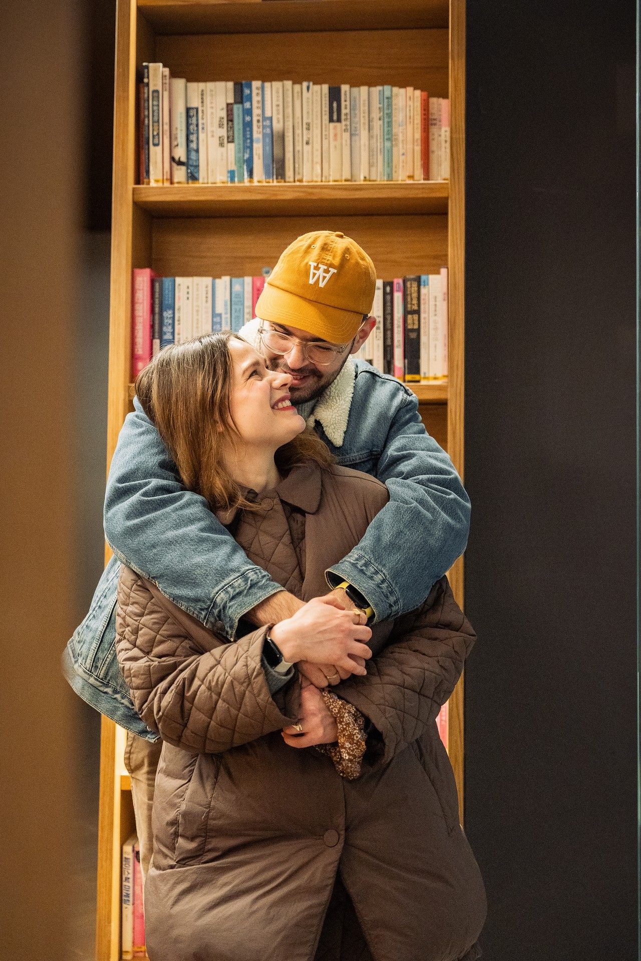 A couple embraces warmly in front of a wooden bookshelf, the man wearing an orange cap and denim jacket.