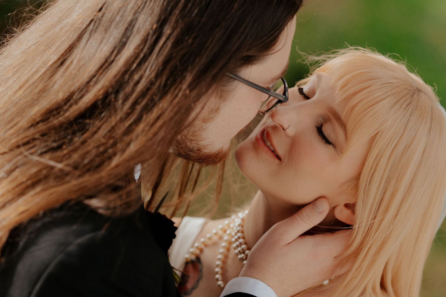 Two people sharing a romantic moment, one with glasses leaning in to kiss a blonde woman wearing pearl necklace outdoors.