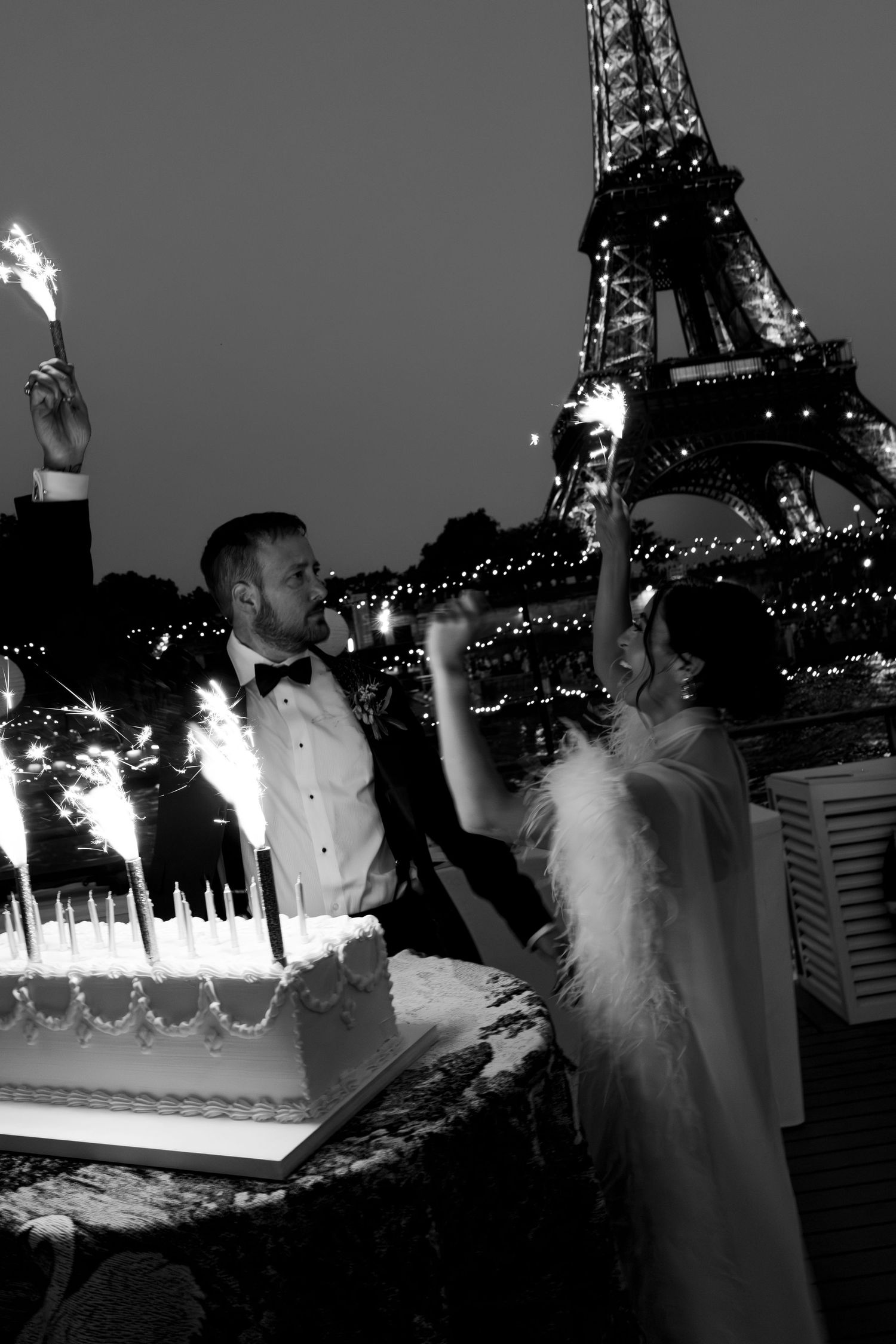 Couple cutting wedding cake with Eiffel Tower lit up behind them in black and white Paris wedding photo. Paris sparklers wedding, film photo