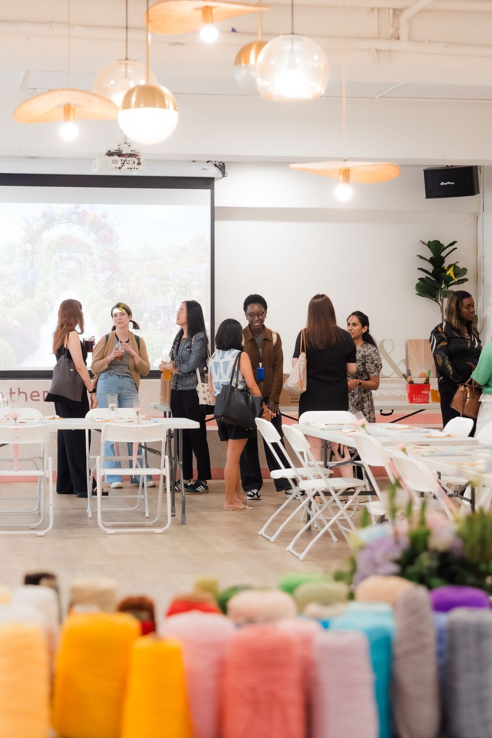 Colorful yarn spools in foreground with diverse women networking at a craft workshop event with projection screen.