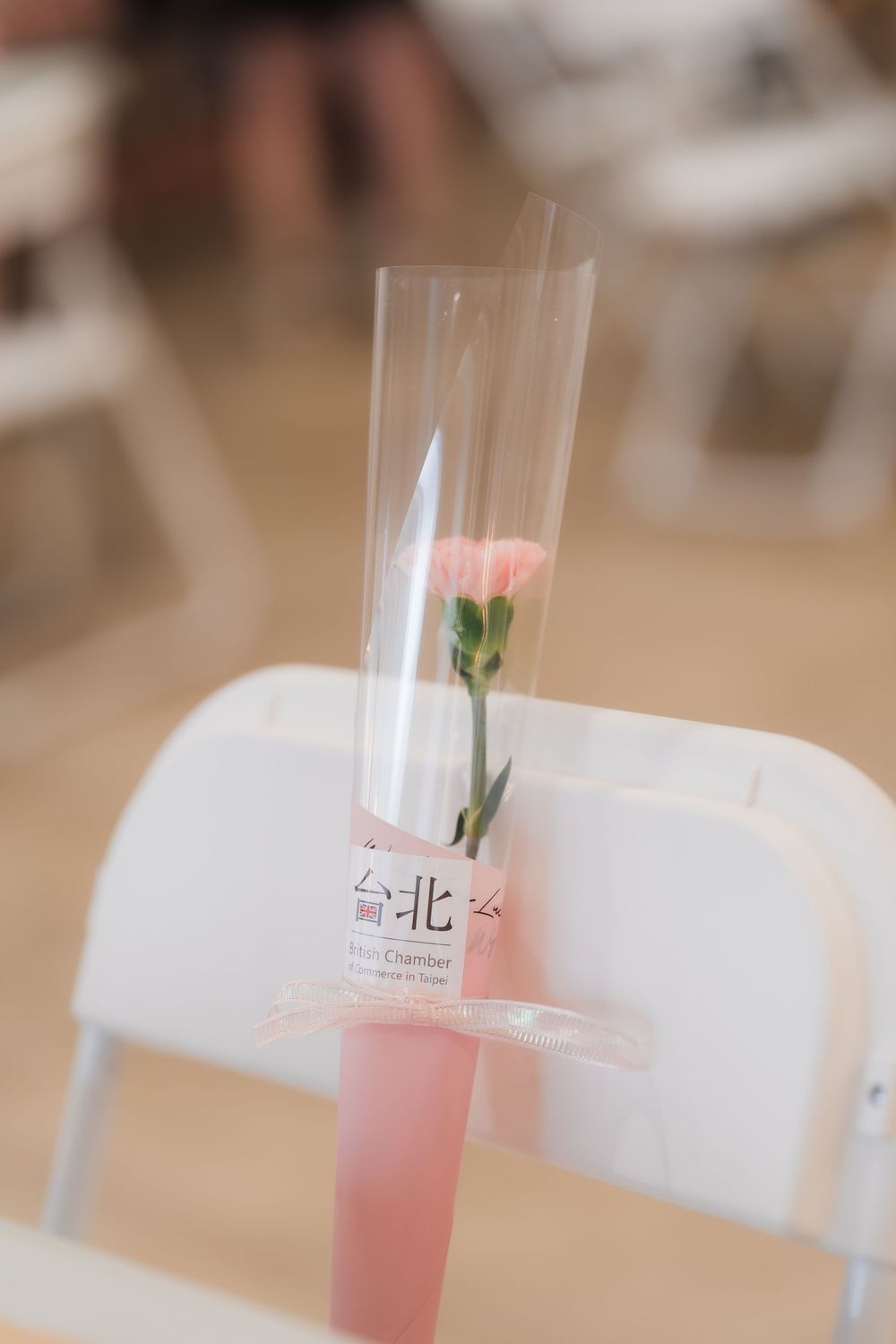A pink carnation flower in a clear cylindrical vase with Chinese text label on a white folding chair.