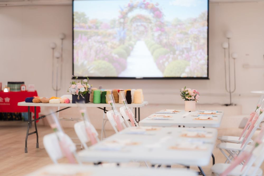 Bright event space with long tables, colorful supplies, and a floral wedding arch displayed on a large projection screen.