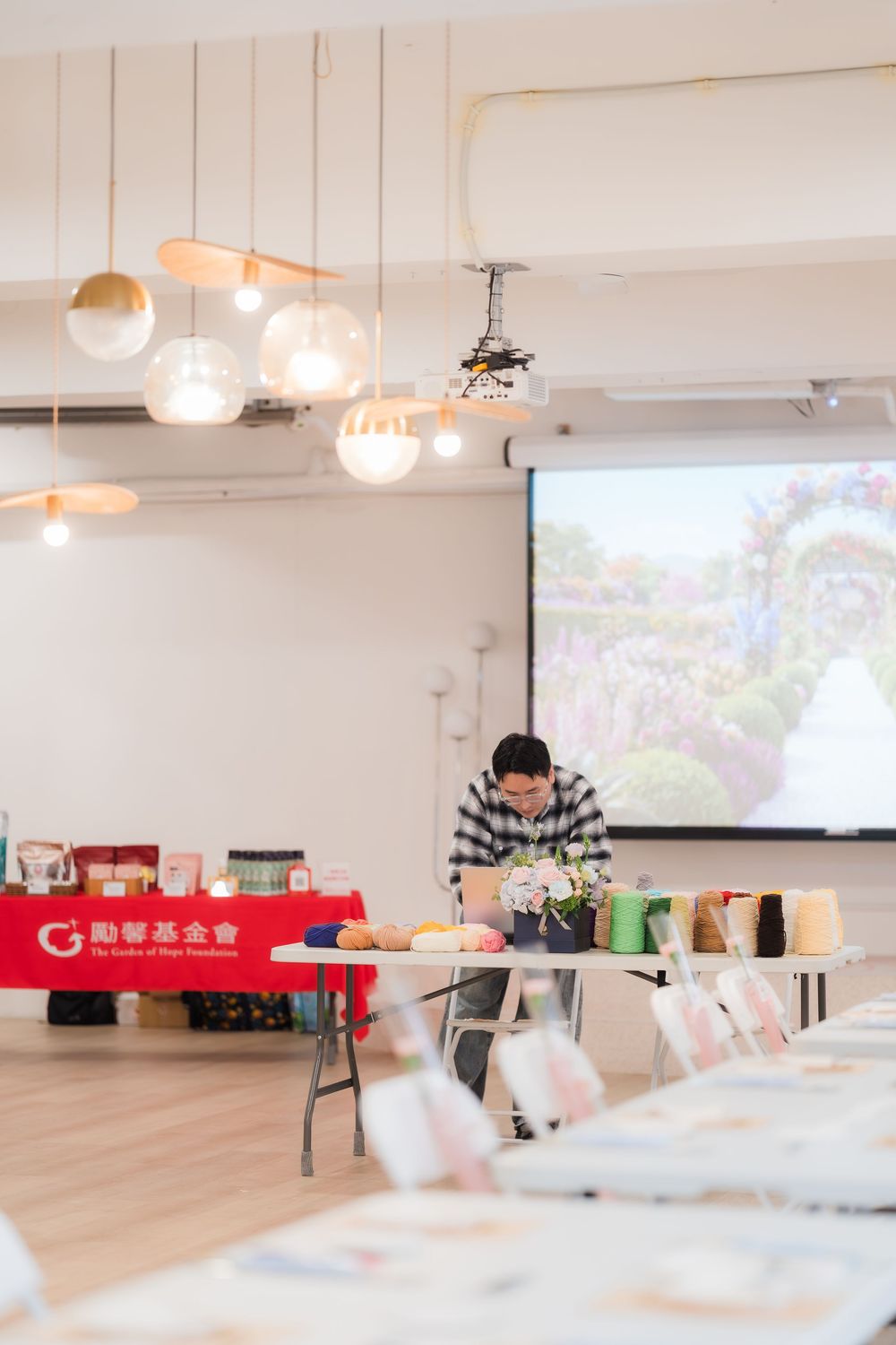 Man arranging floral displays at a workshop table with a garden image projected on screen behind him.