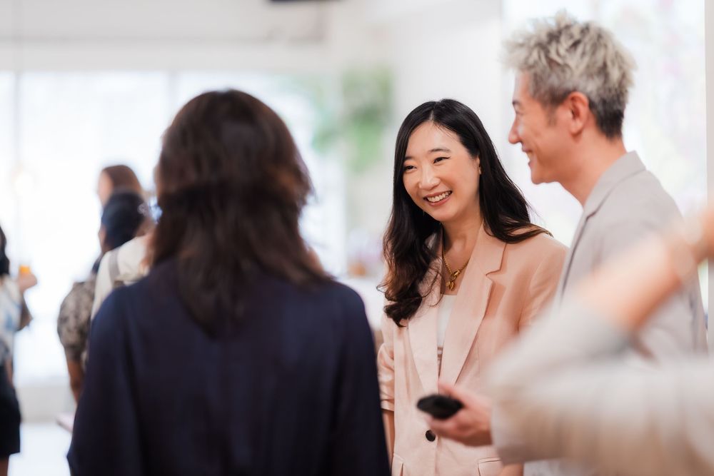 Smiling Asian woman in beige blazer engaging in conversation at a professional networking event.