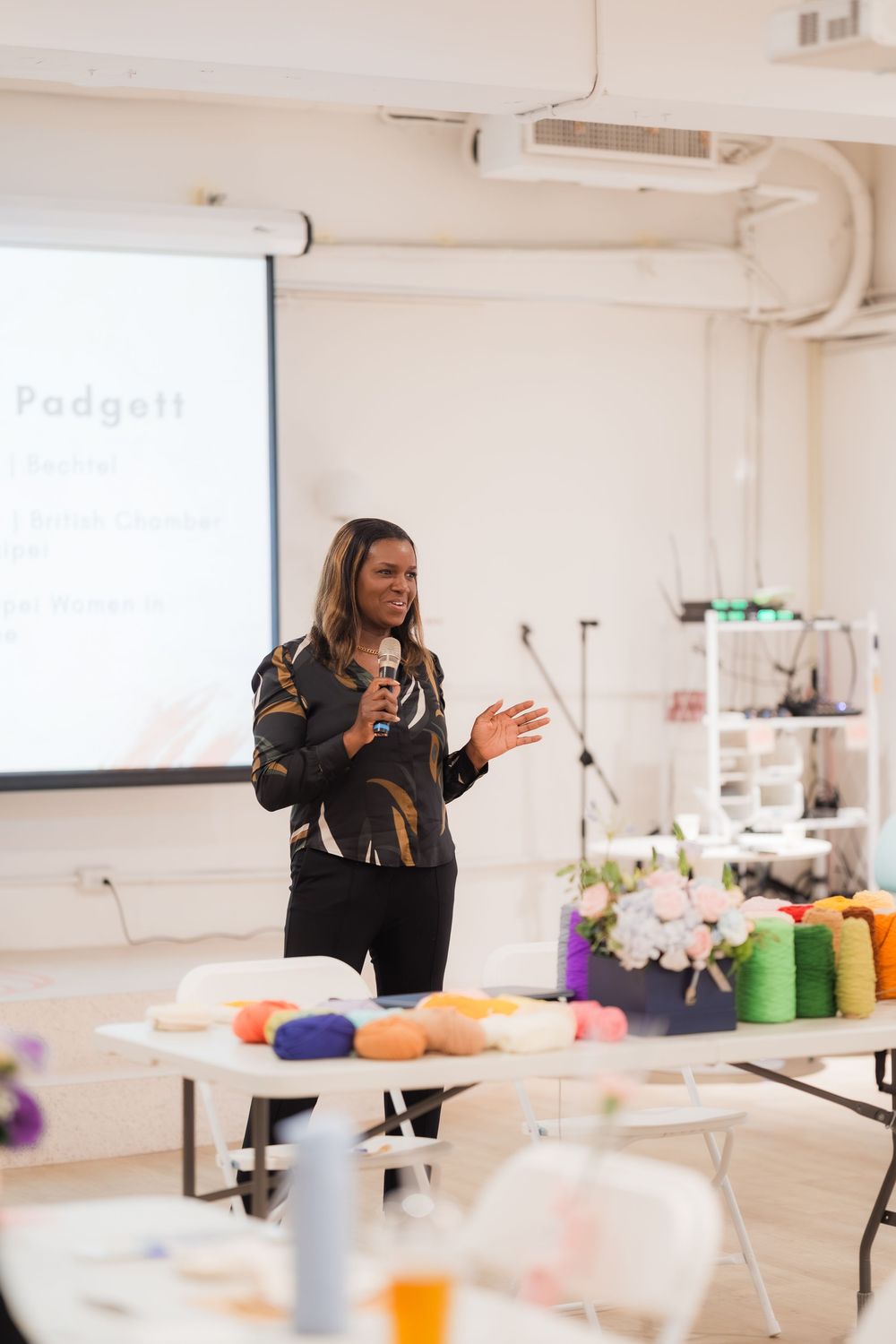 Woman speaker holds microphone presenting at a workshop with colorful yarn and craft supplies on tables.