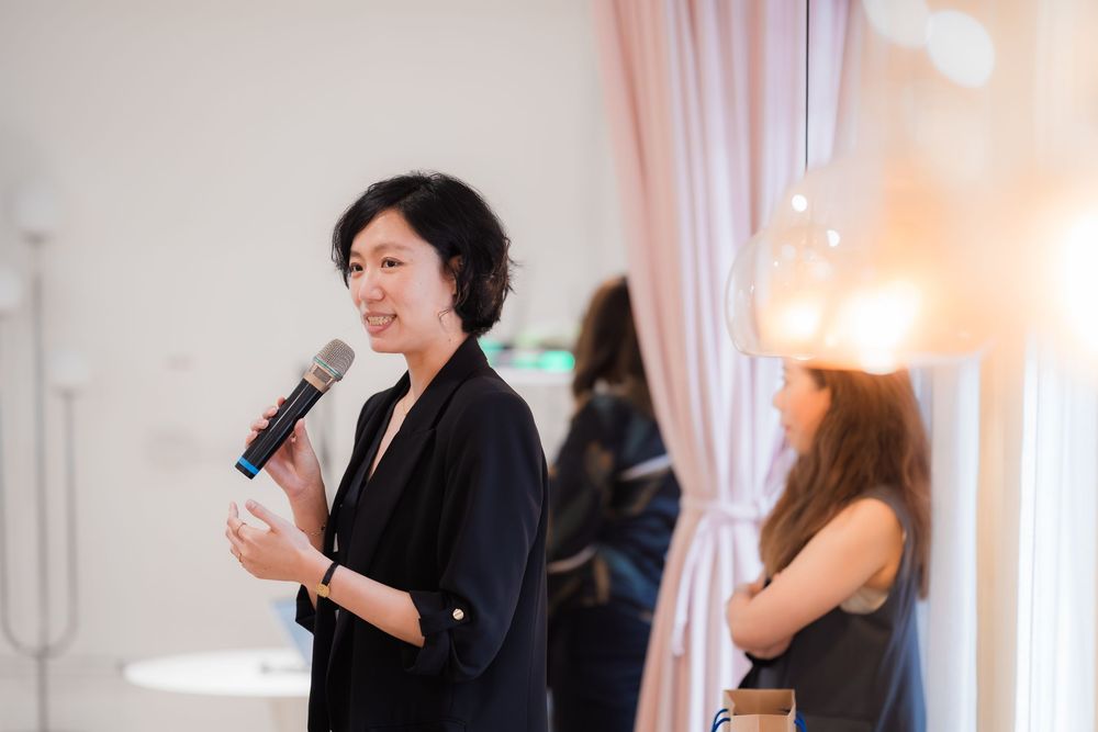Asian woman speaking into microphone at indoor event, wearing black blazer, with soft lighting and guests in background.