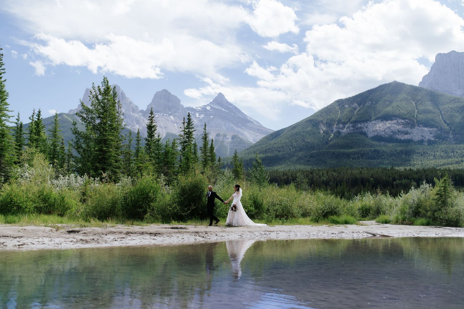 Bride and groom pose by a turquoise river with stunning mountain backdrop in Canmore, Alberta wedding photo.