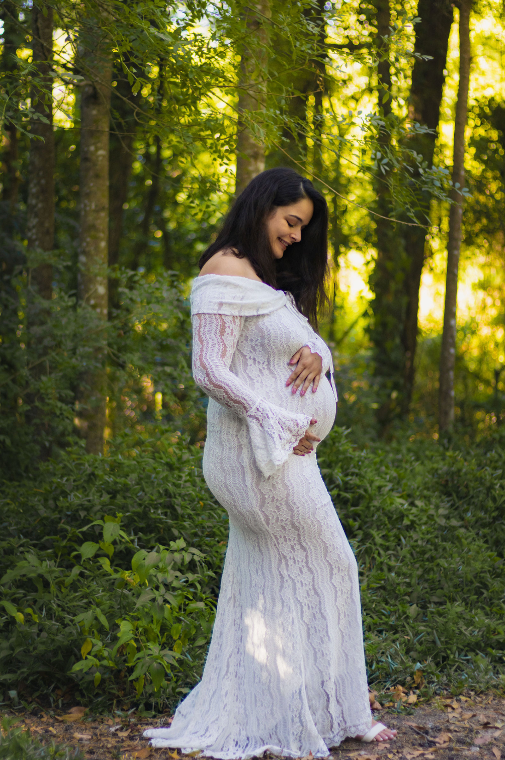 Pregnant woman in white lace off-shoulder gown cradling baby bump in sunlit forest setting.