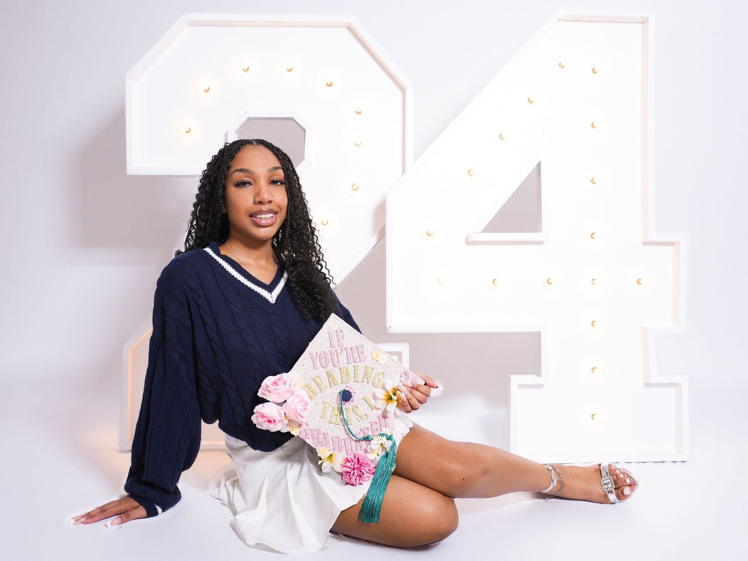 Young woman in navy sweater poses with decorated graduation cap in front of large illuminated '24' for Class of 2024.