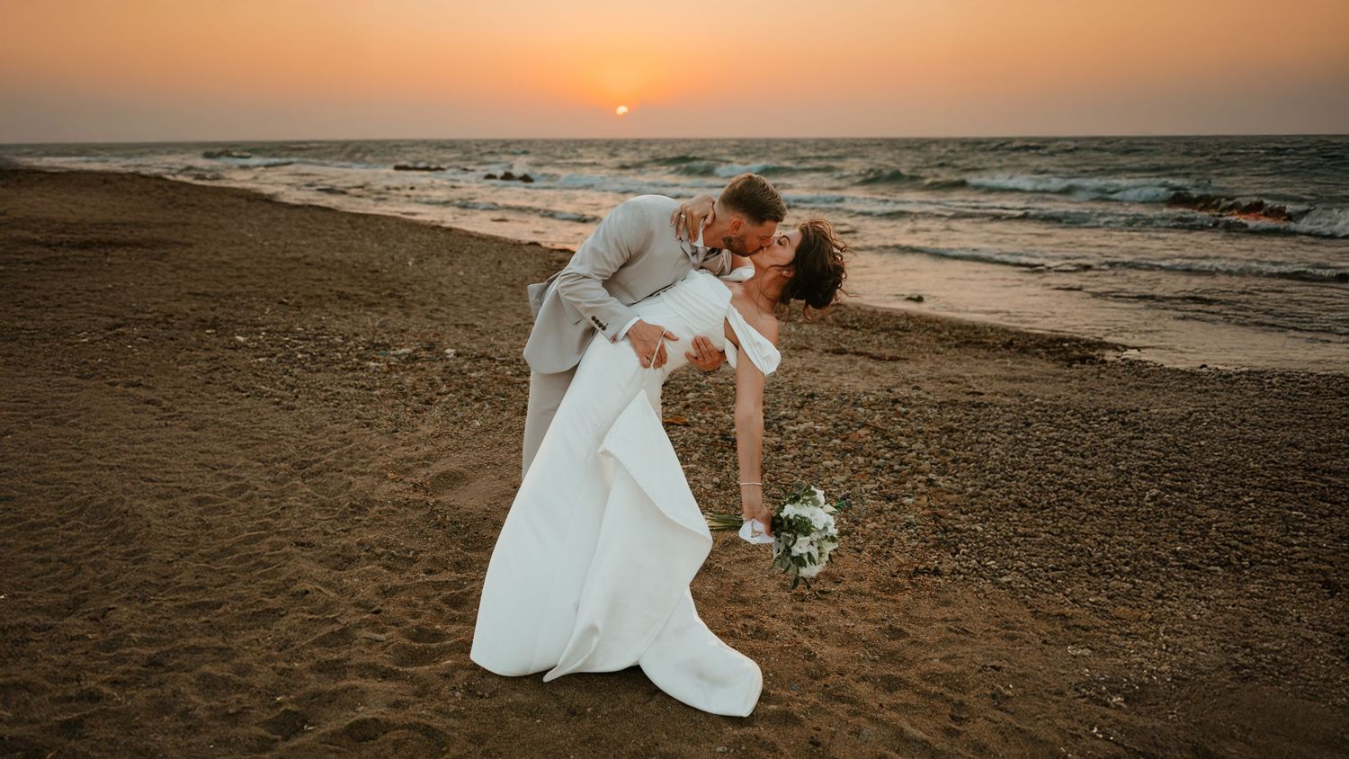 Bride and groom share a romantic dip kiss on a sandy beach at sunset with ocean waves in the background.