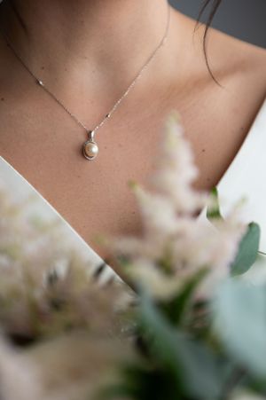 Close-up view of a delicate silver necklace with a pearl pendant against blurred floral background.