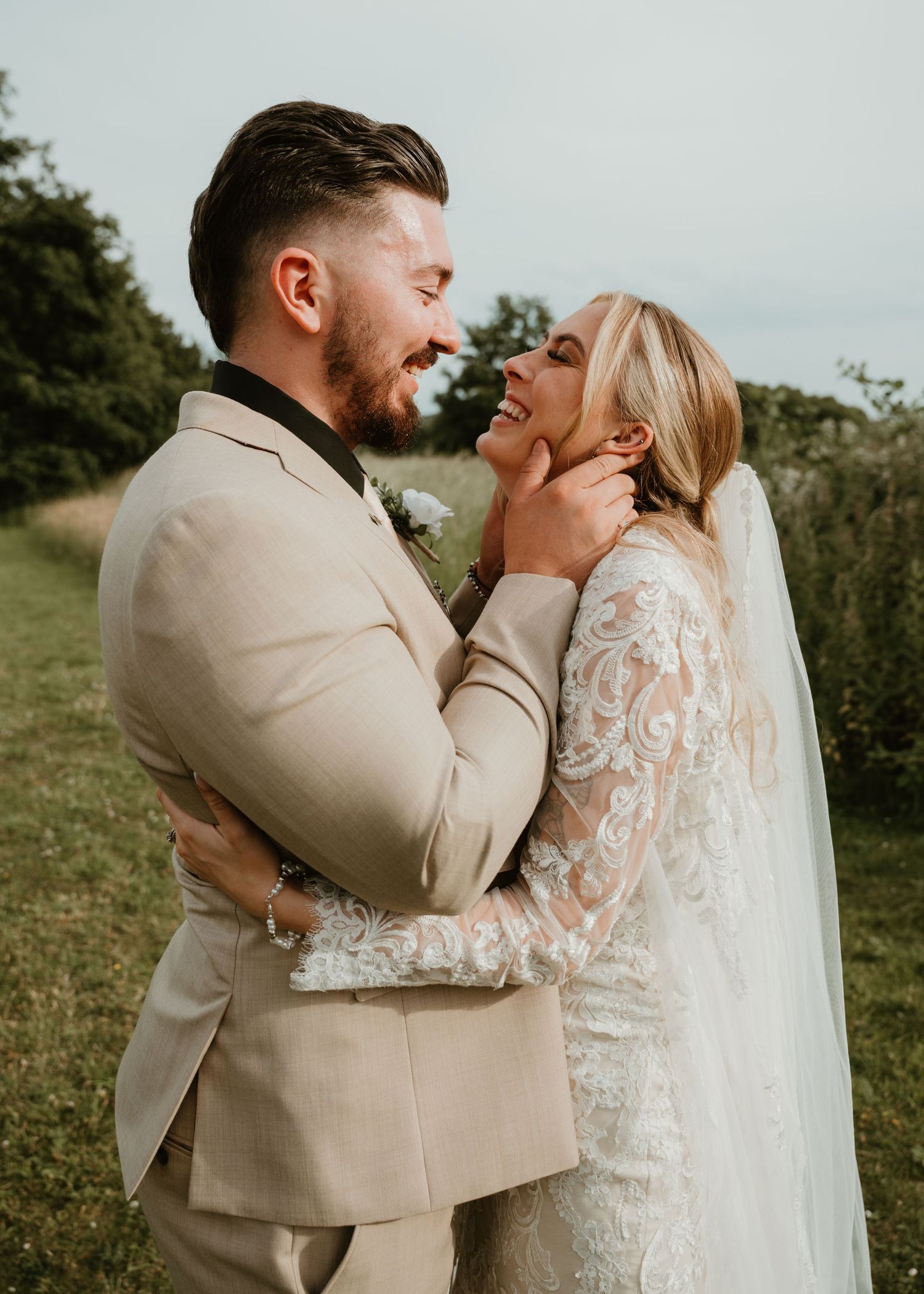 A smiling bride in lace gown and groom in beige suit share a tender moment outdoors on their wedding day.