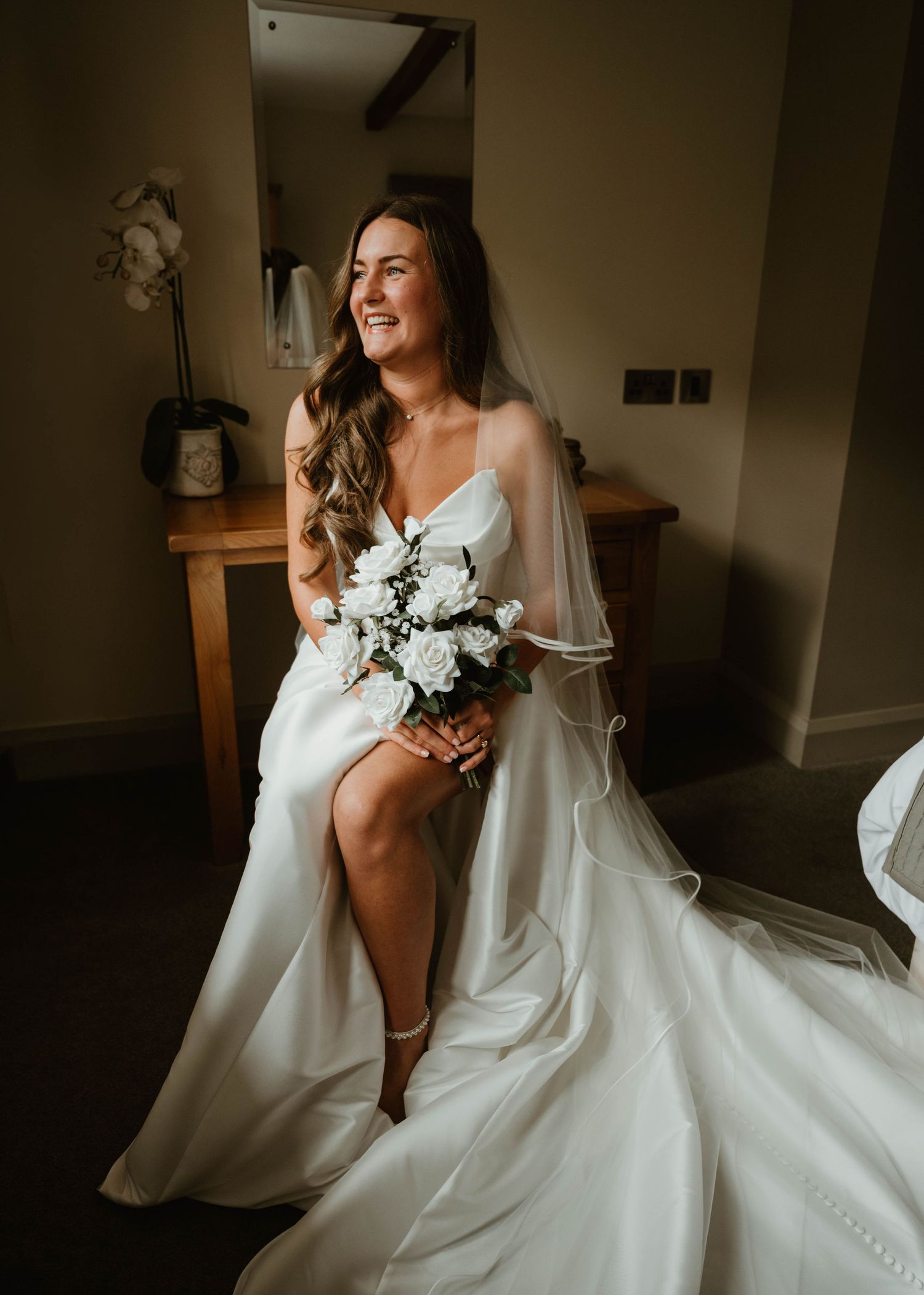 A radiant bride in a white satin gown with veil holds a white rose bouquet, smiling joyfully in a warmly lit room.
