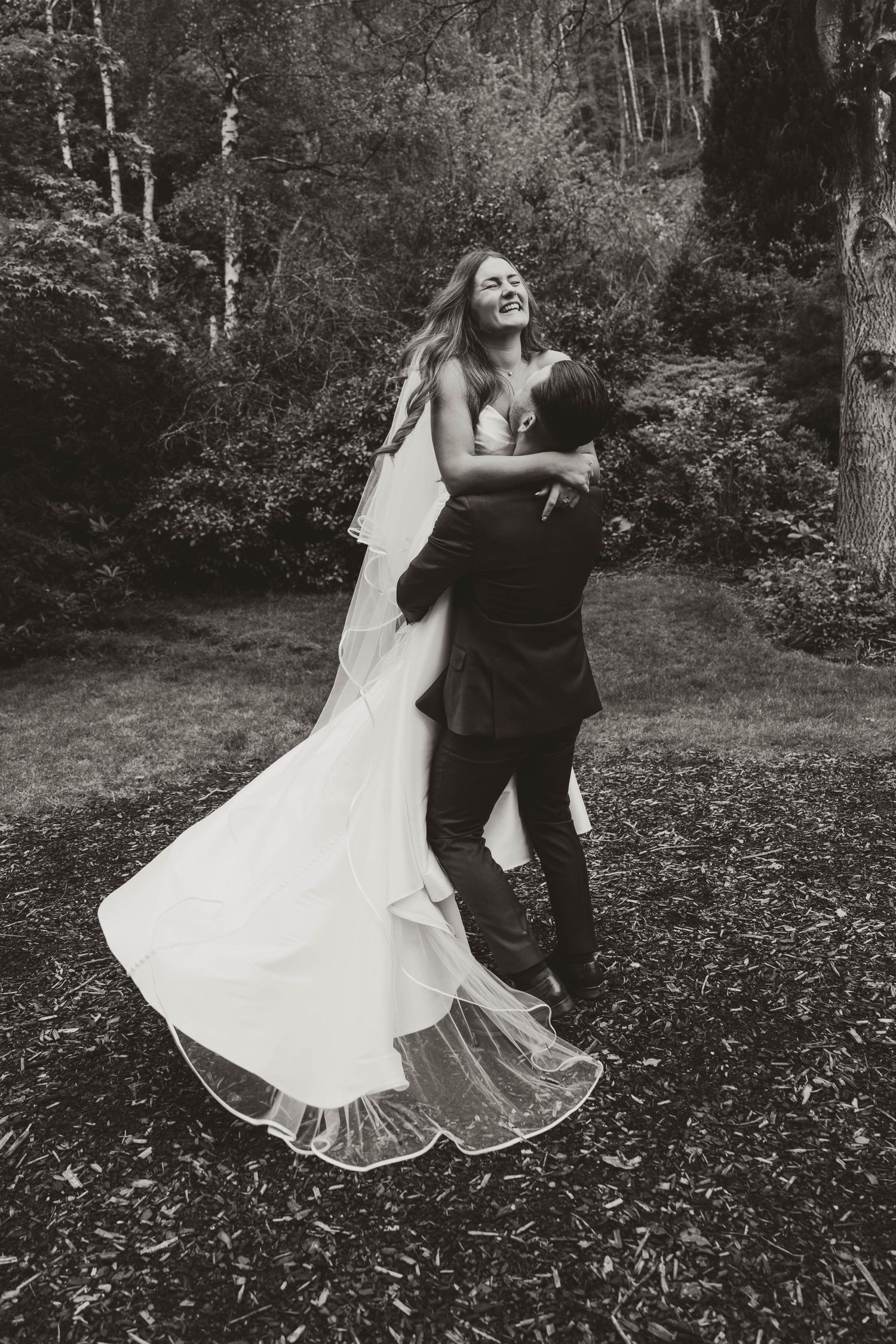 Black and white photo of a groom lifting his laughing bride in a wedding dress outdoors.
