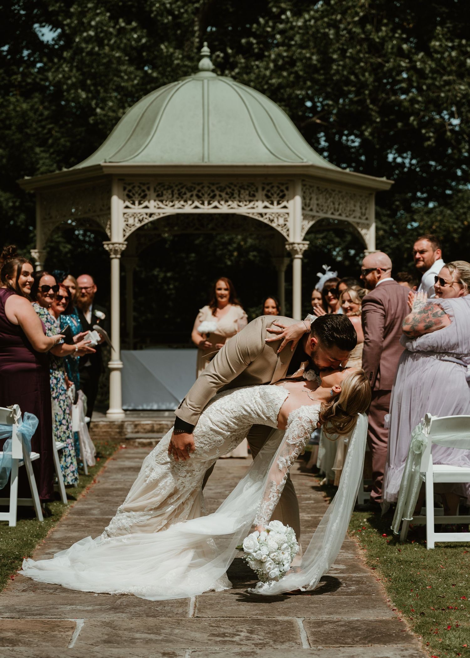 Groom dips bride for a romantic kiss at outdoor wedding ceremony near elegant white gazebo, surrounded by guests.