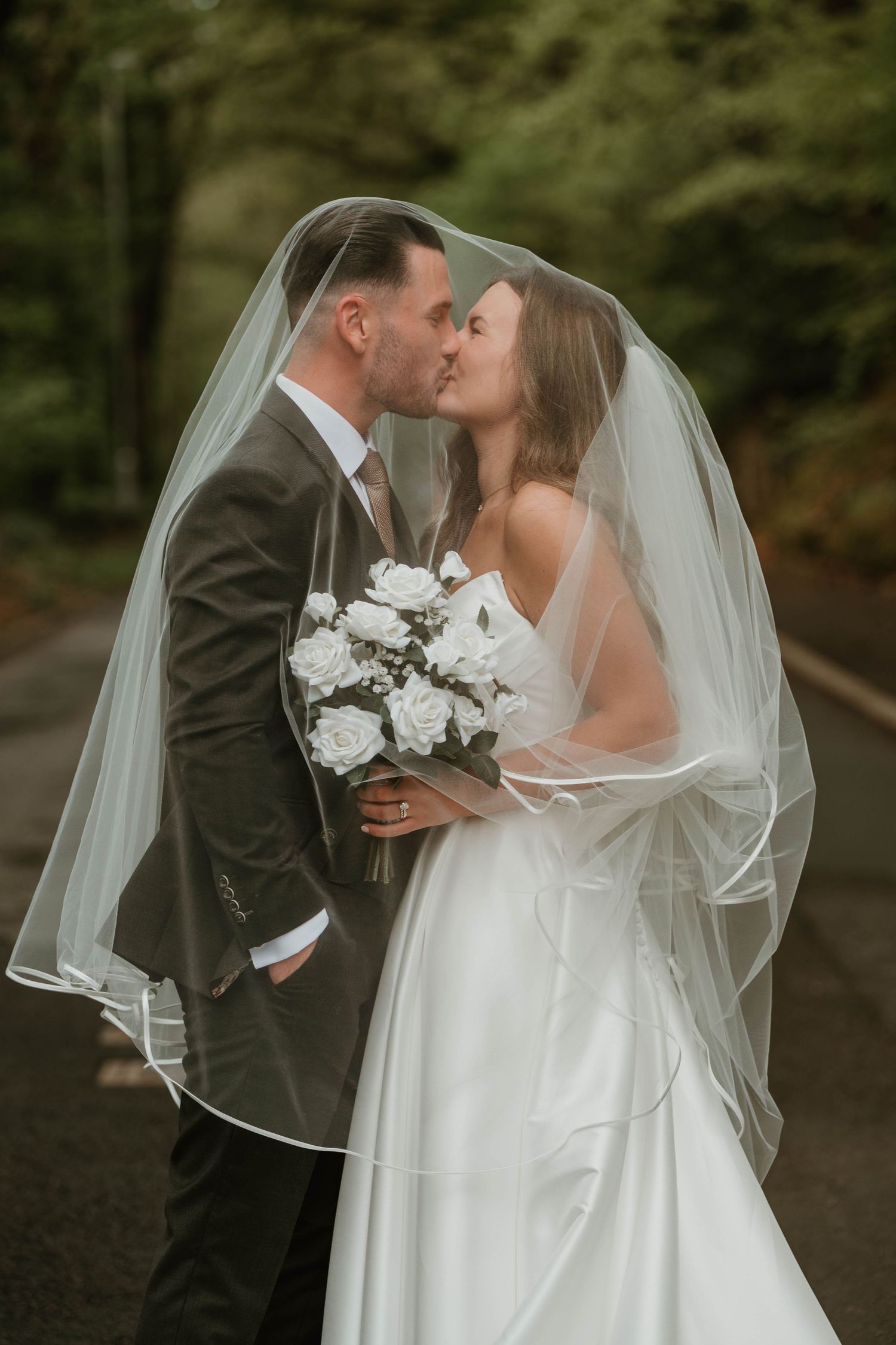Bride and groom kissing under a wedding veil, bride holding white rose bouquet, surrounded by lush green trees.