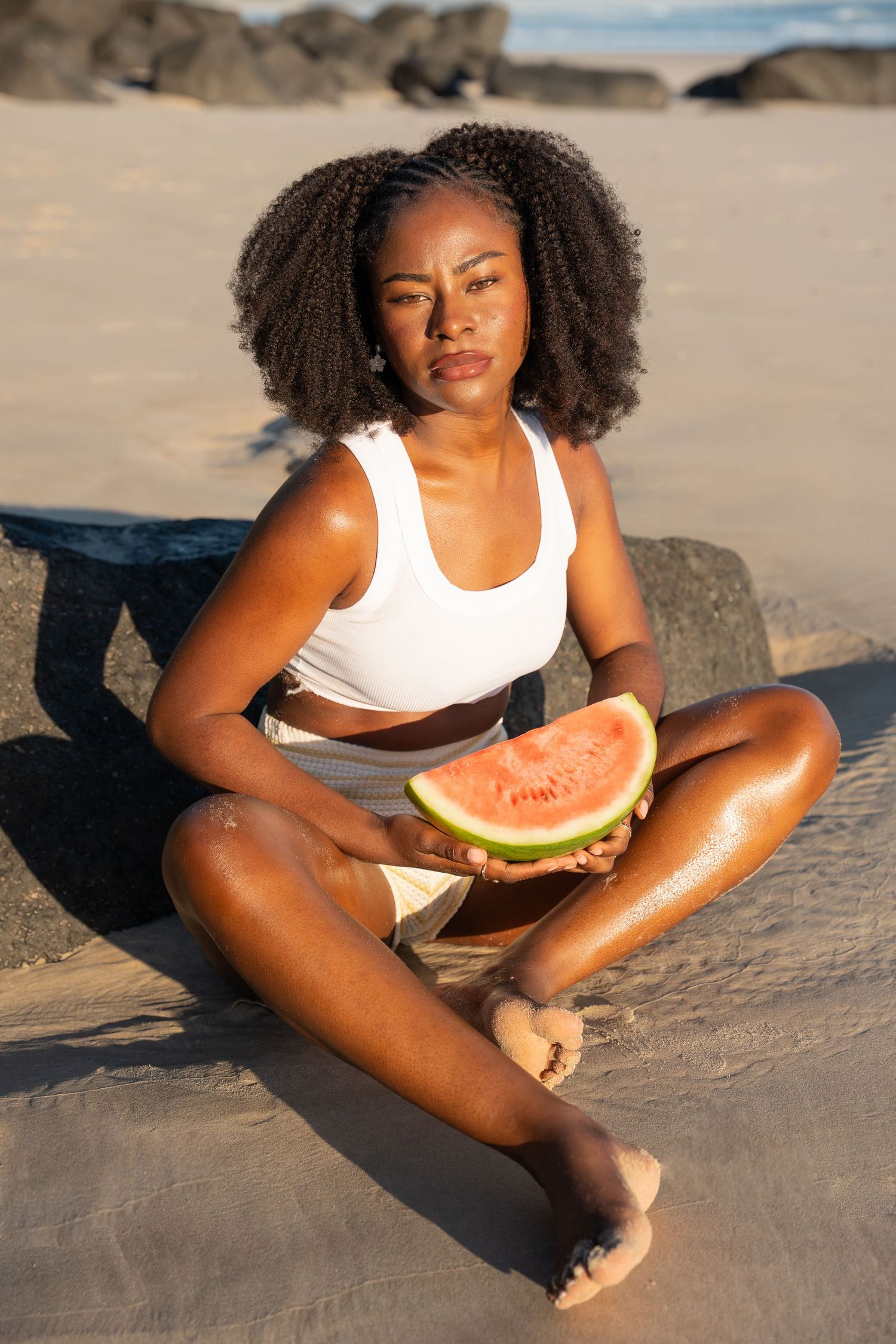 Young Black woman with curly hair sits cross-legged on sandy beach holding a fresh watermelon slice in golden sunlight.
