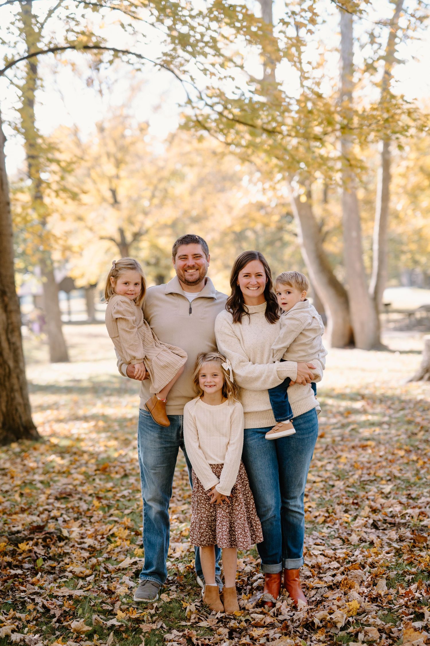 Family of five posing outdoors in autumn park with golden fall foliage, wearing coordinated neutral and cream outfits.