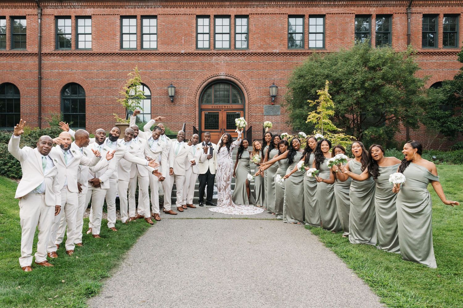 Wedding party poses outside brick venue, groomsmen in white suits and bridesmaids in sage green gowns holding white bouquets.