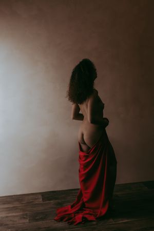 Woman with curly hair draped in red silk fabric posing against a neutral wall in artistic fine art photography.
