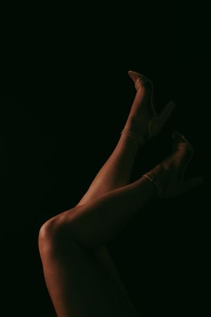 Elegant female legs in clear heels posed against a dark black background in dramatic low-key lighting.