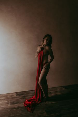 A woman poses nude holding a flowing red fabric draped elegantly in a dimly lit studio with wooden floors.