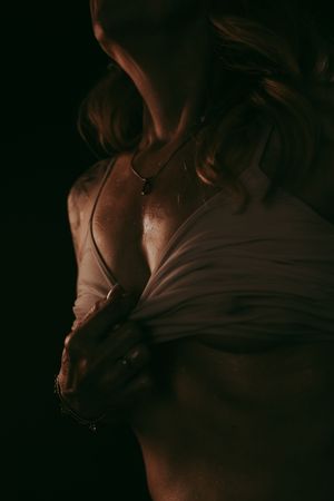 A woman in soft dramatic lighting holds a white fabric, wearing a delicate necklace against a dark background.