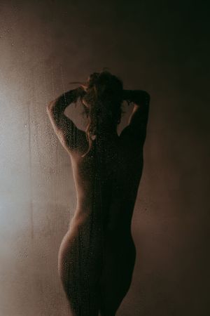 Silhouette of a woman showering behind a wet glass door with moody, dramatic lighting.
