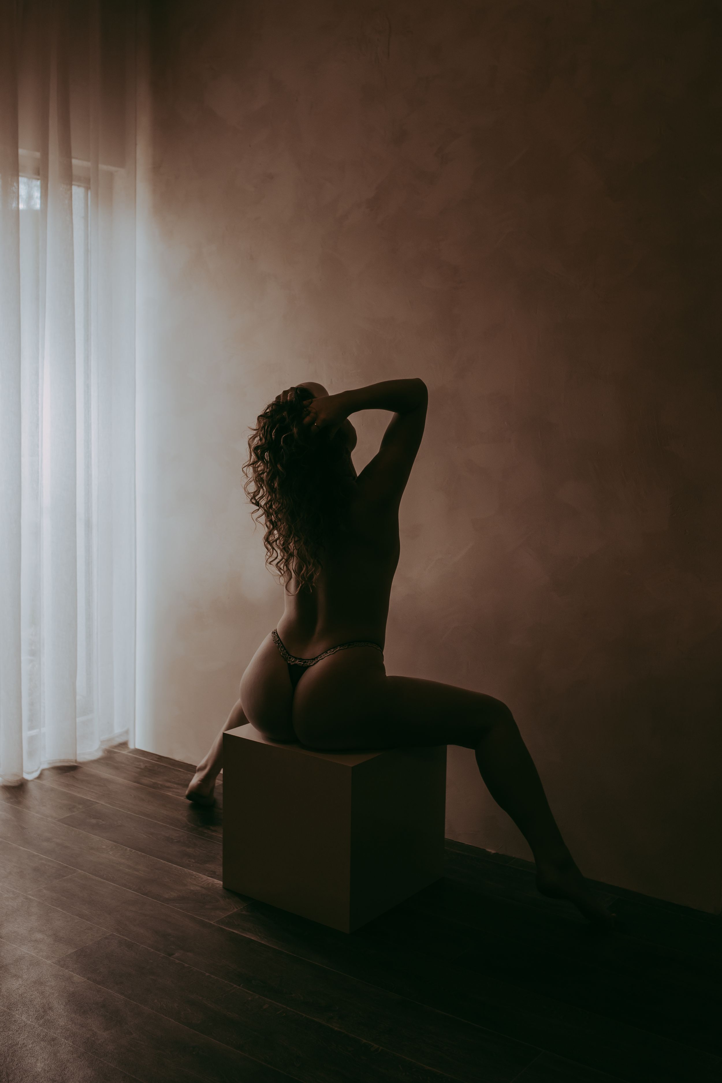 Silhouette of a woman in lingerie sitting on a cube near a curtained window in a dimly lit room.