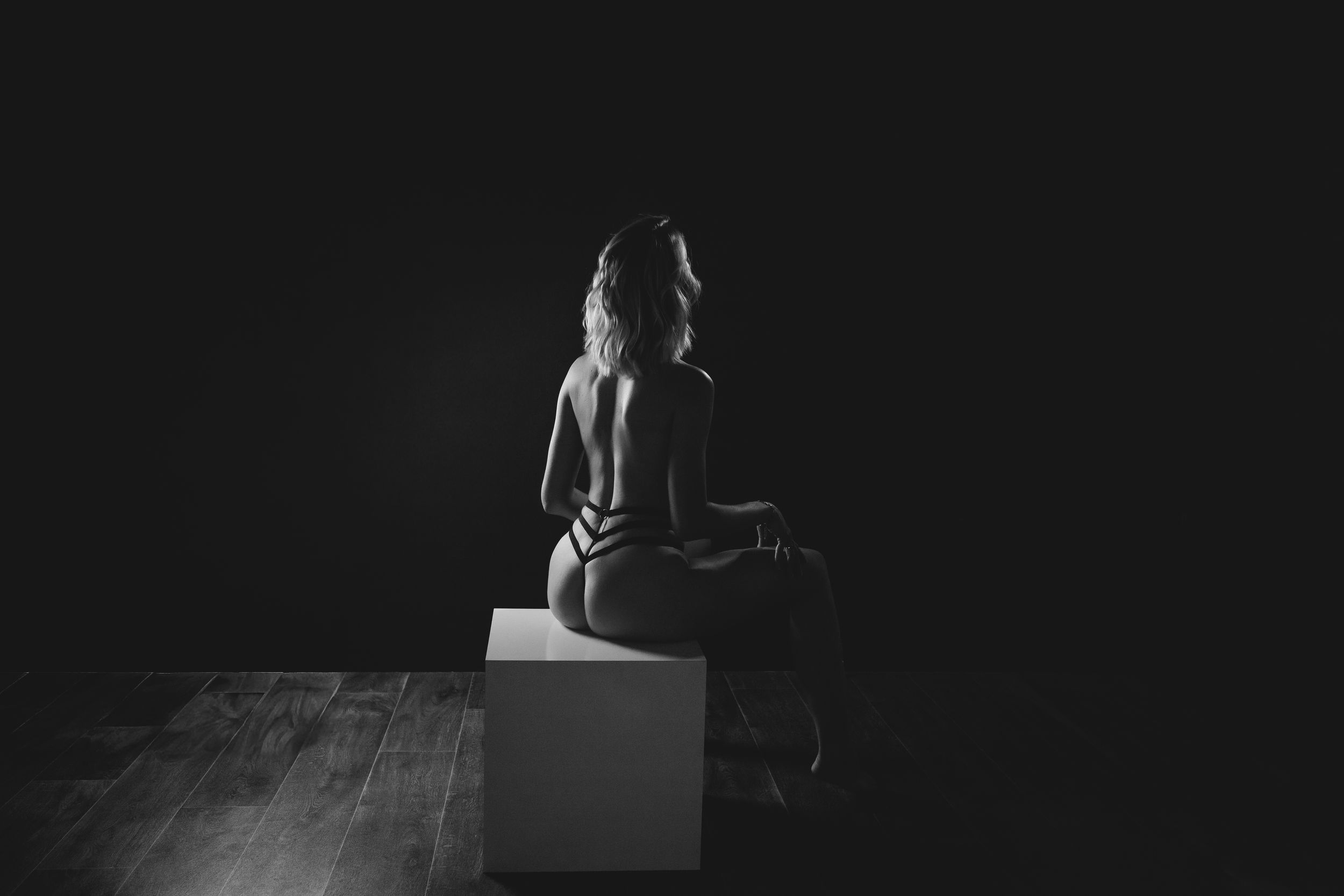 Black and white fine art photo of a woman sitting on a white cube in a dark studio, viewed from behind.