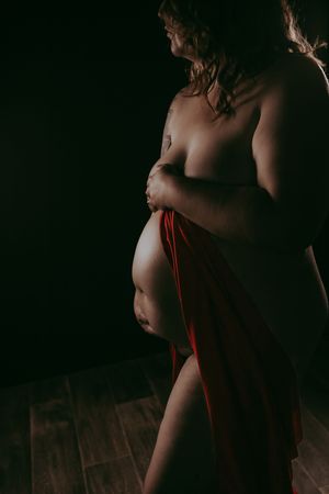 Artistic maternity photo of pregnant woman draped in red fabric against dark background, cradling bare belly.